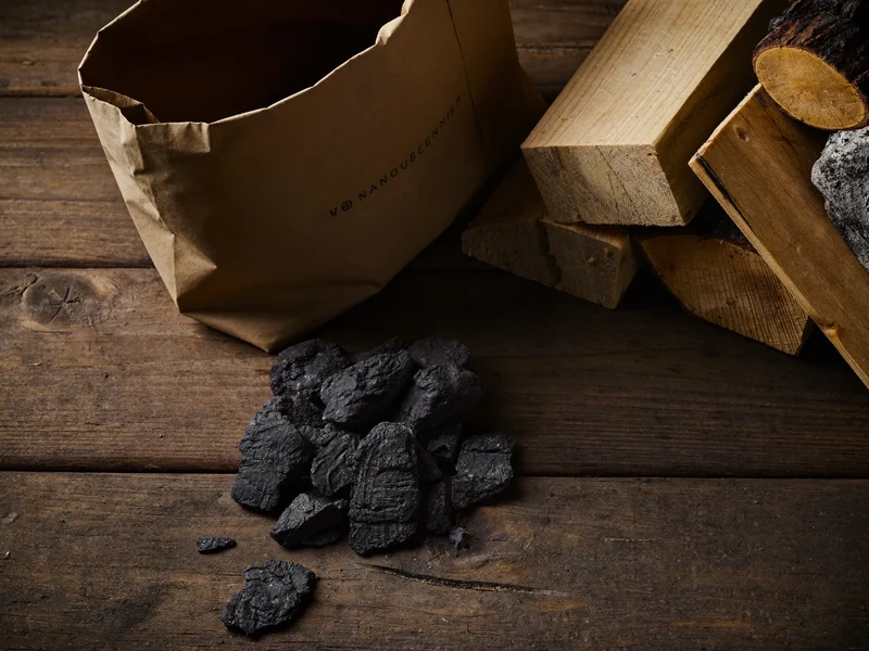 Papiersack mit Naturholzkohle neben Kohlestücken und Buchenholz auf Holztisch