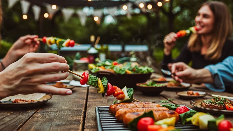 Hände am Gartentisch bei Dämmerung mit Würstchen und Gemüsespießen auf dem Rost